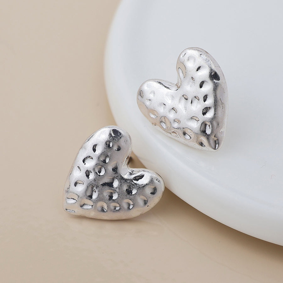 Pair of silver-plated, hammered-texture heart earrings resting on a white ceramic surface.