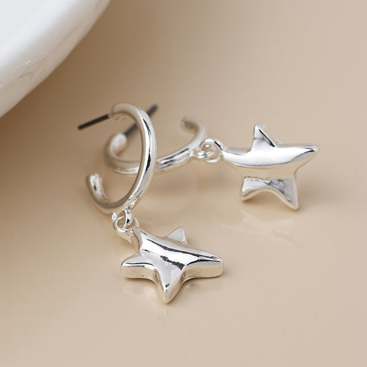 A pair of silver-plated C-hoop earrings, each featuring a dangling, three-dimensional star charm, displayed on a light beige surface.