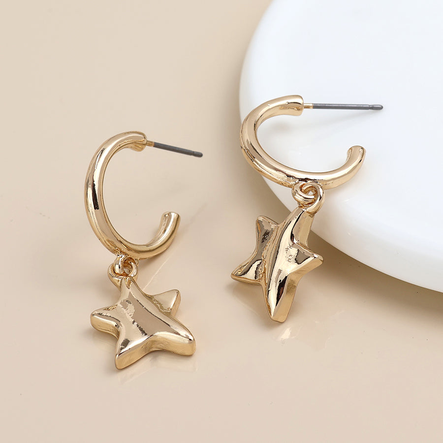 A pair of gold C-hoop earrings with hanging star-shaped charms, displayed on a beige surface.