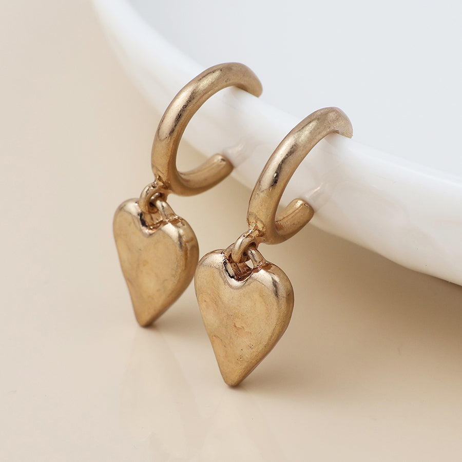 A pair of small gold earrings with dangling heart-shaped charms, evoking a vintage style, displayed beside a white dish.