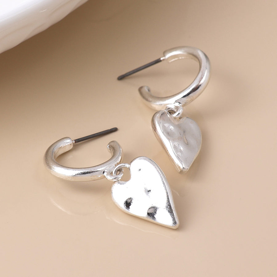 A pair of vintage silver earrings featuring a hoop design and dangling, slightly textured heart-shaped charms rests on a beige surface.