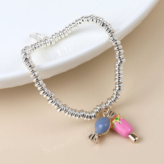 A silver-plated charm bracelet featuring a pink and blue sweet-shaped charm sits on a light surface beside a white dish, capturing the playful spirit of confectionery-inspired jewellery.