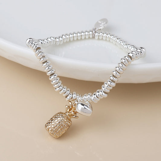 A silver-plated beaded stretch bracelet featuring a gold pineapple charm and a small silver oval charm, displayed on a white dish.