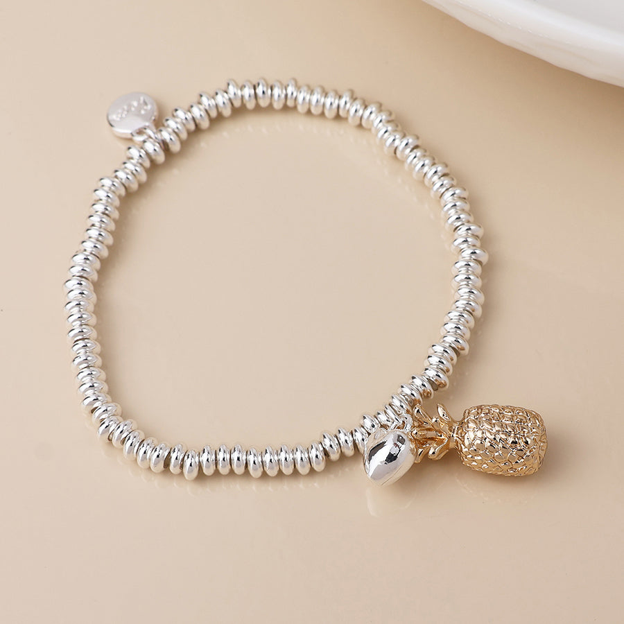 A stretch bracelet with silver-plated beads showcases two charms—a gleaming metallic oval and a gold pineapple—presented on a beige surface.