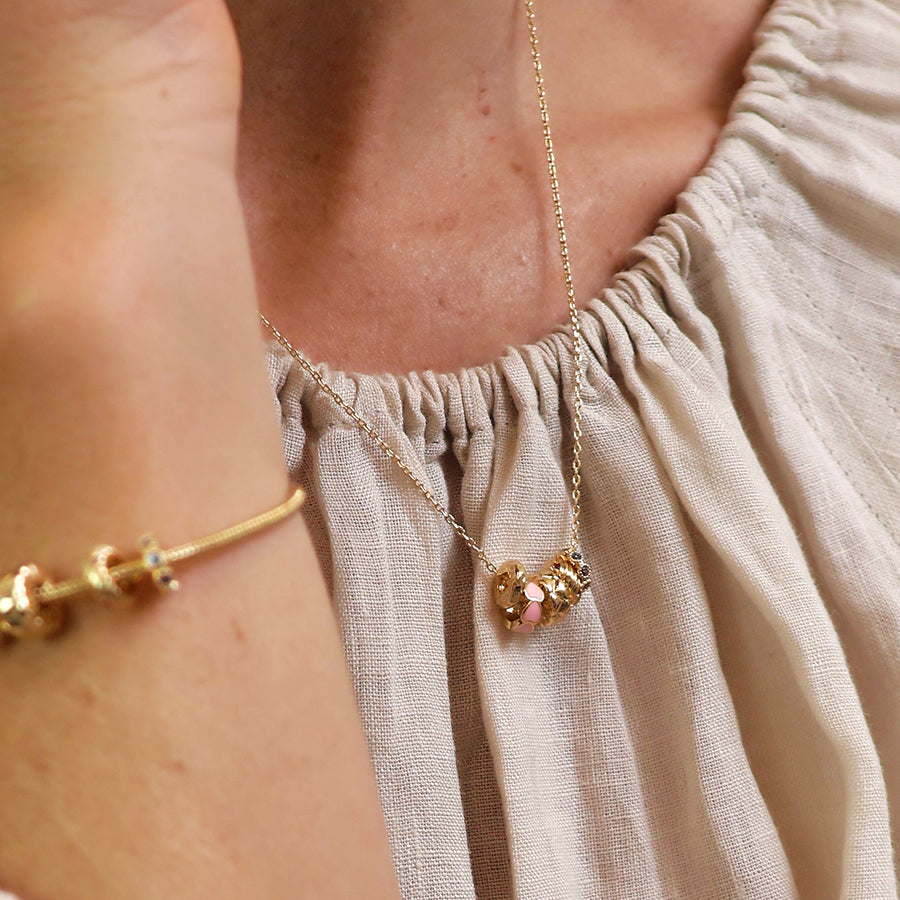 A person wearing a light, textured top and a gold hoop necklace featuring a heart-shaped pendant, accompanied by a gold bracelet on their wrist.