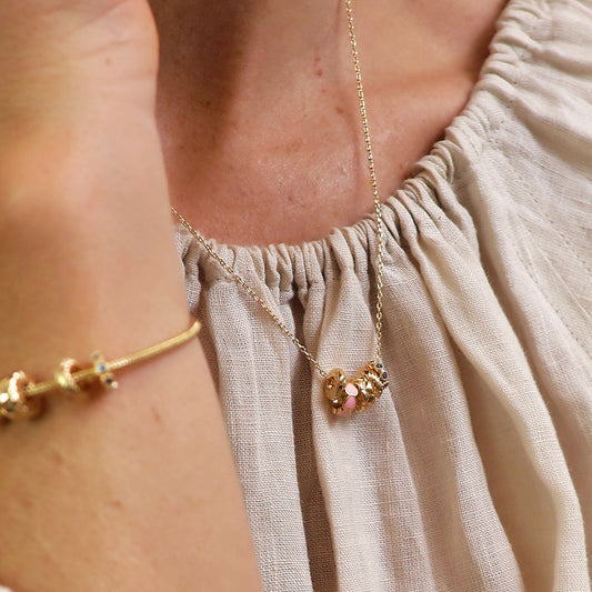 A person wearing a light, textured top and a gold hoop necklace featuring a heart-shaped pendant, accompanied by a gold bracelet on their wrist.
