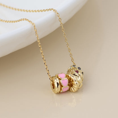Gold chain necklace featuring three gold charms, including a pink heart accent and a floral motif, displayed on a light-coloured surface.
