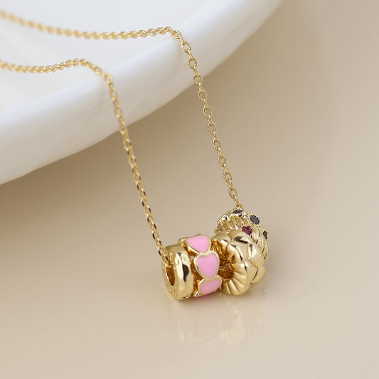 Gold chain necklace featuring three gold charms, including a pink heart accent and a floral motif, displayed on a light-coloured surface.