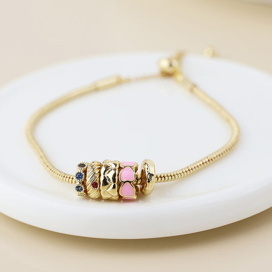 A golden hoop bracelet decorated with pink hearts and multicoloured crystals, featuring gold, pink, and blue accents, presented on a white circular plate.