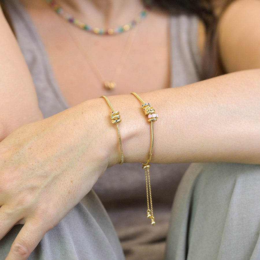 Individual wearing two gold adjustable bracelets adorned with small charms and pink hearts on their wrist, paired with a colourful beaded necklace, seated with arms resting on their knees.