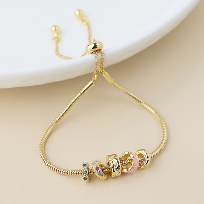 A gold hoop bracelet adorned with five decorative charms, including pink hearts and multicoloured crystals, shown on a light surface beside a white dish.