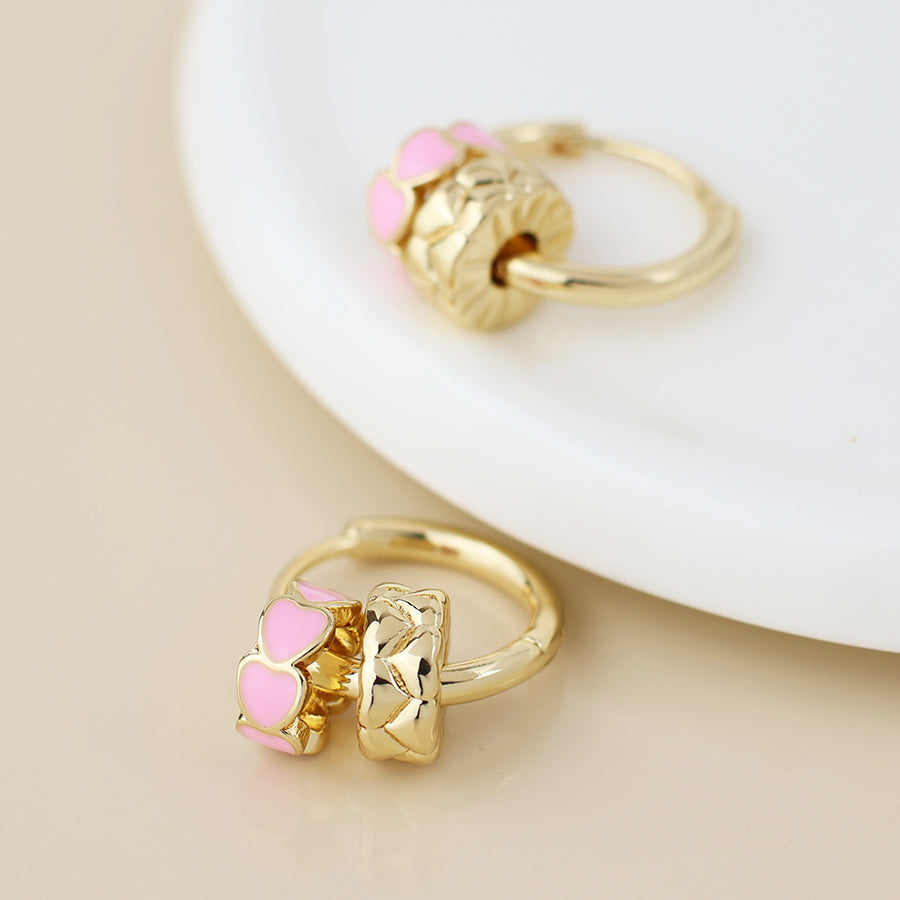 Two gold-tone earrings, each featuring a gold charm and a pink enamel heart-shaped charm, are displayed on a white ceramic surface.