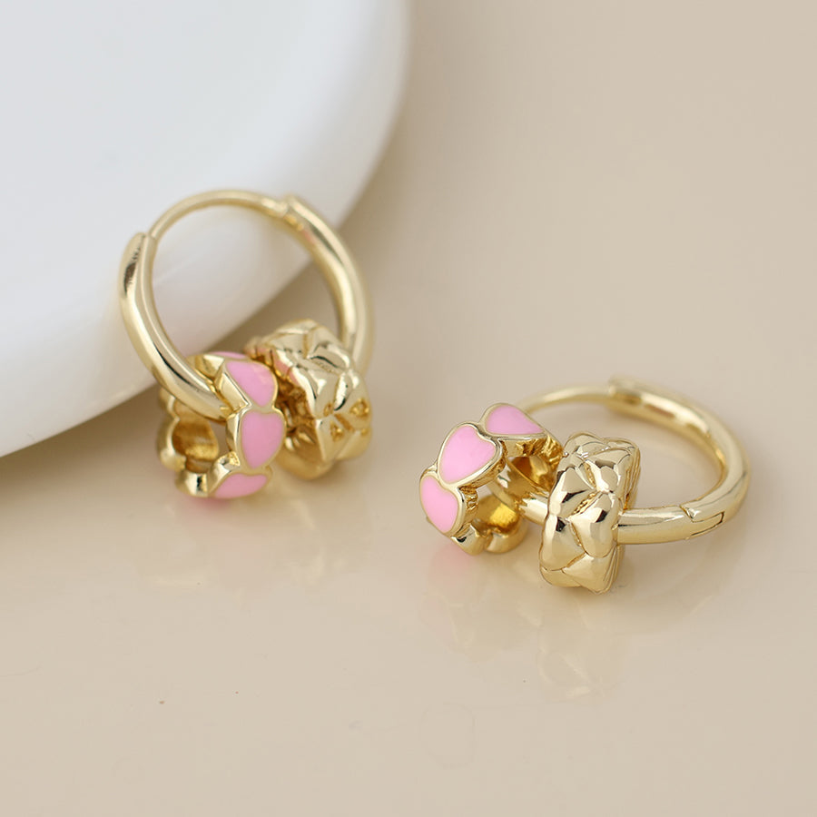 A pair of gold-tone, heart-shaped hoop earrings with gold and pink enamel detailing, displayed on a beige surface.