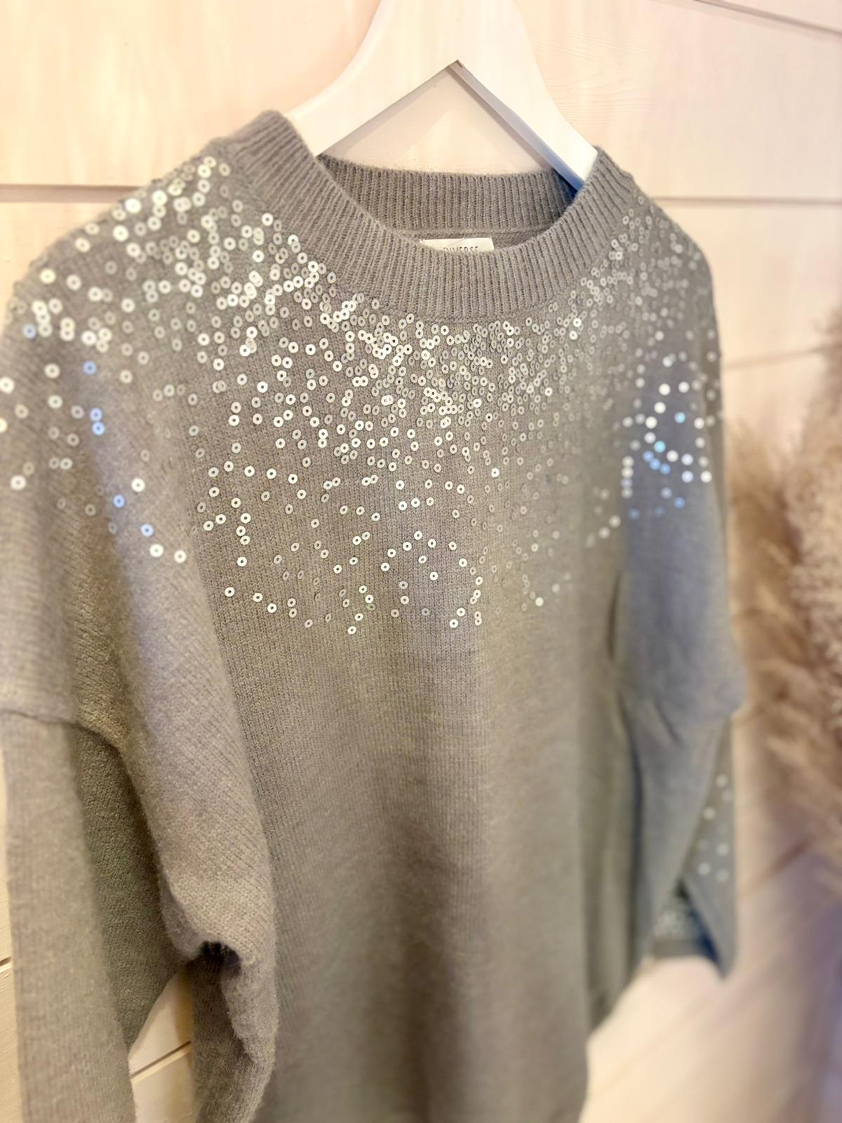 A grey sequin jumper adorned with silver sequins scattered across the upper front and sleeves hangs on a white hanger—ideal for layering, set against a pale wooden wall.