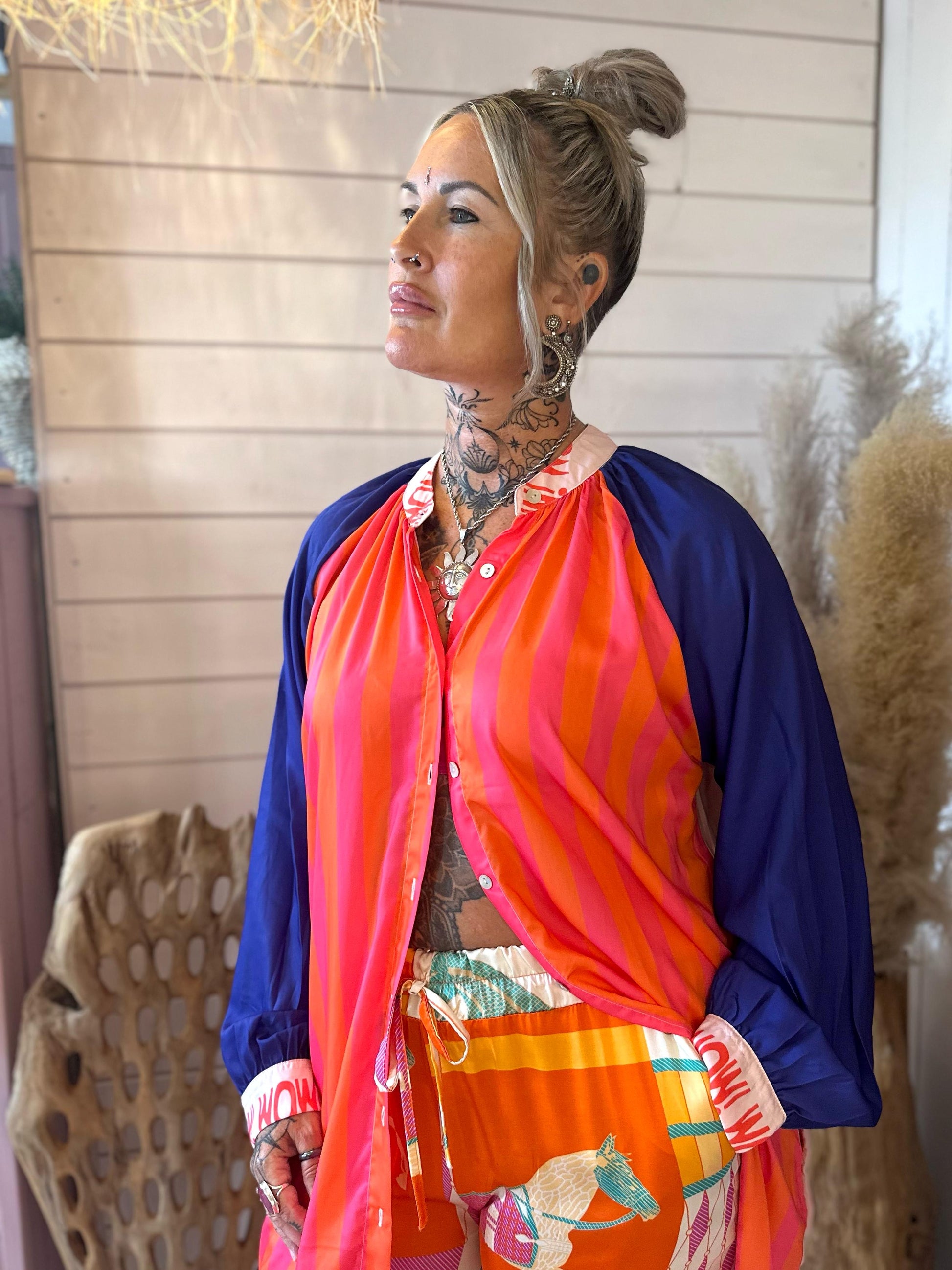 A woman with tattoos stands indoors, wearing a colourful outfit with a pink and orange striped top and patterned shorts, looking off to the side.