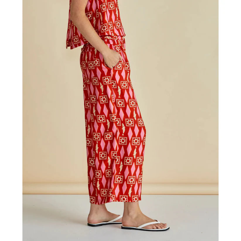 Person in red patterned lightweight trousers and flip-flops, standing side-on with one hand in a pocket against a plain beige background—highlighting relaxed, ethical fashion.