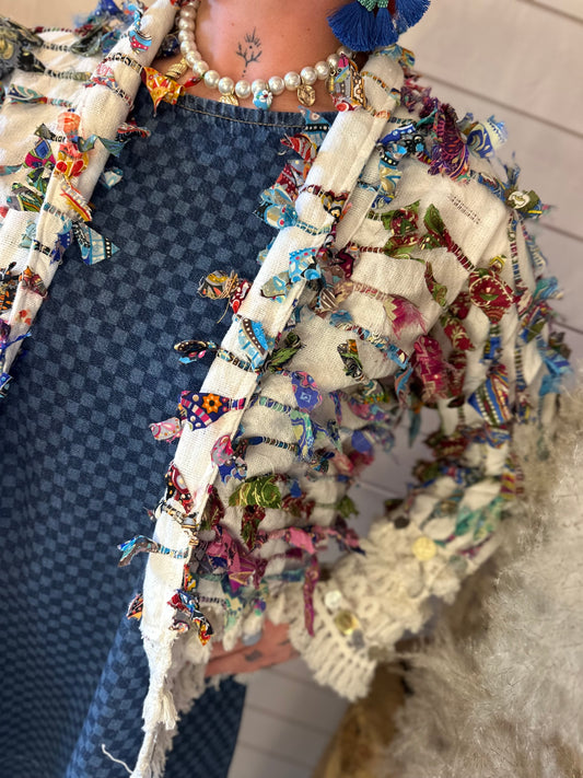 A person wearing a blue checked dress, a hippy jacket with colourful fabric knots, a pearl necklace, and a blue tassel earring—perfect for festival season—stands in front of a light wooden background.