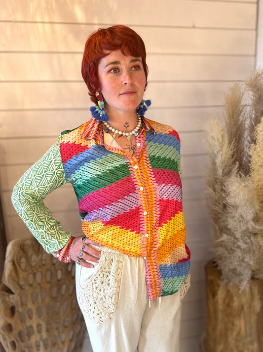 A person with short red hair wears a funky statement shirt with colourful stripes, white trousers with lace pockets, and beaded jewellery, standing in front of a light wooden wall and pampas grass—perfect for festival season.