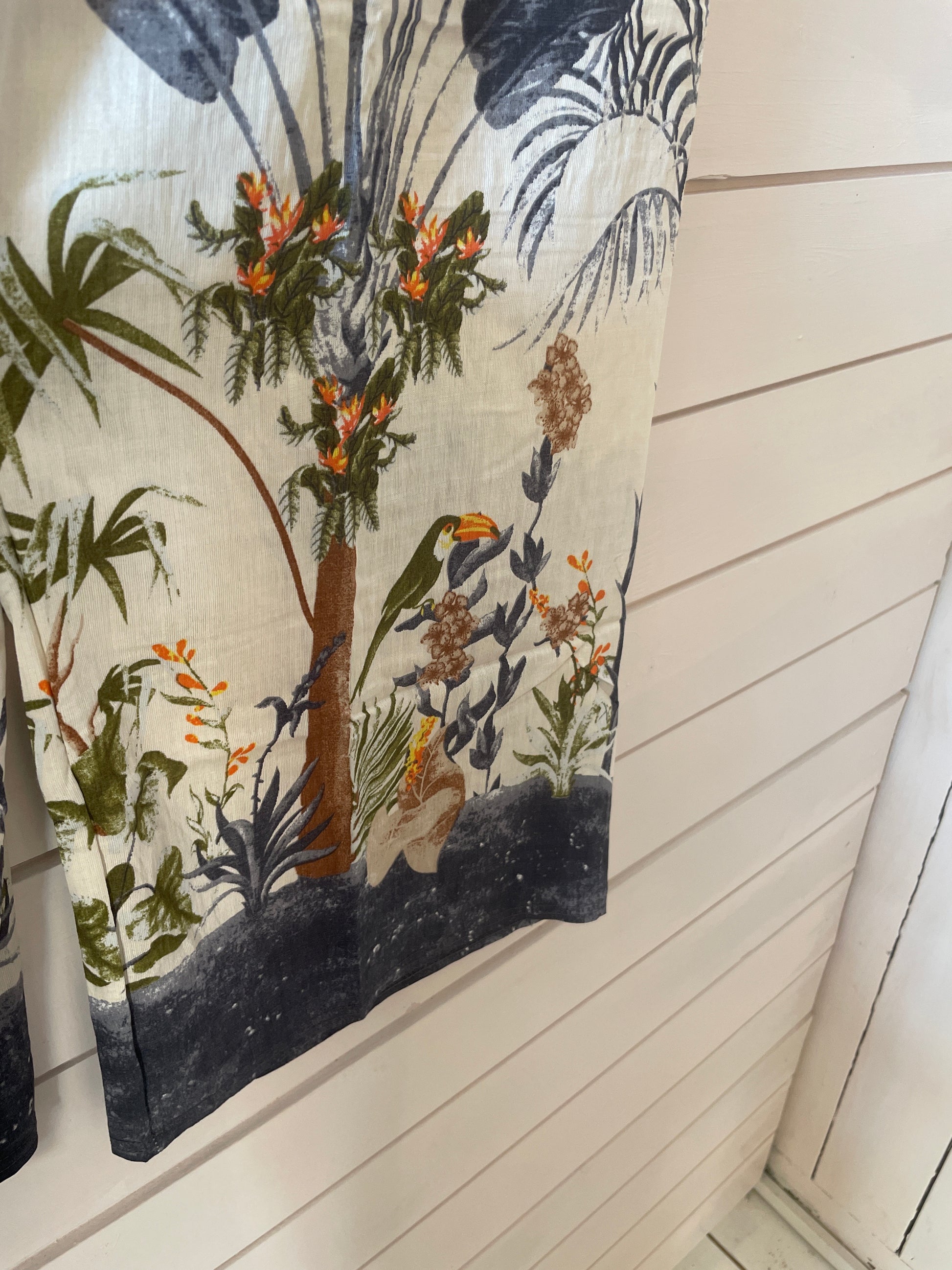 Fabric with a tropical print showing a toucan, flowers, and foliage hangs against a white wooden wall—ideal for sewing blue jungle trousers with an elasticated waistband.