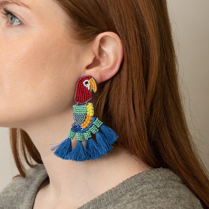 A woman wearing a large, colourful parrot earring shaped like a beaded parrot with blue fringe at the bottom.