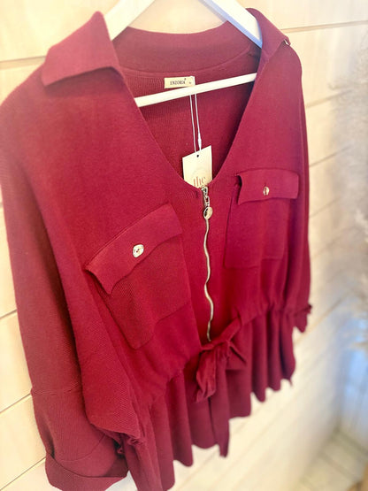 A maroon, buttoned blouse with front pockets and a zip, shown on a white hanger against a light wooden wall.