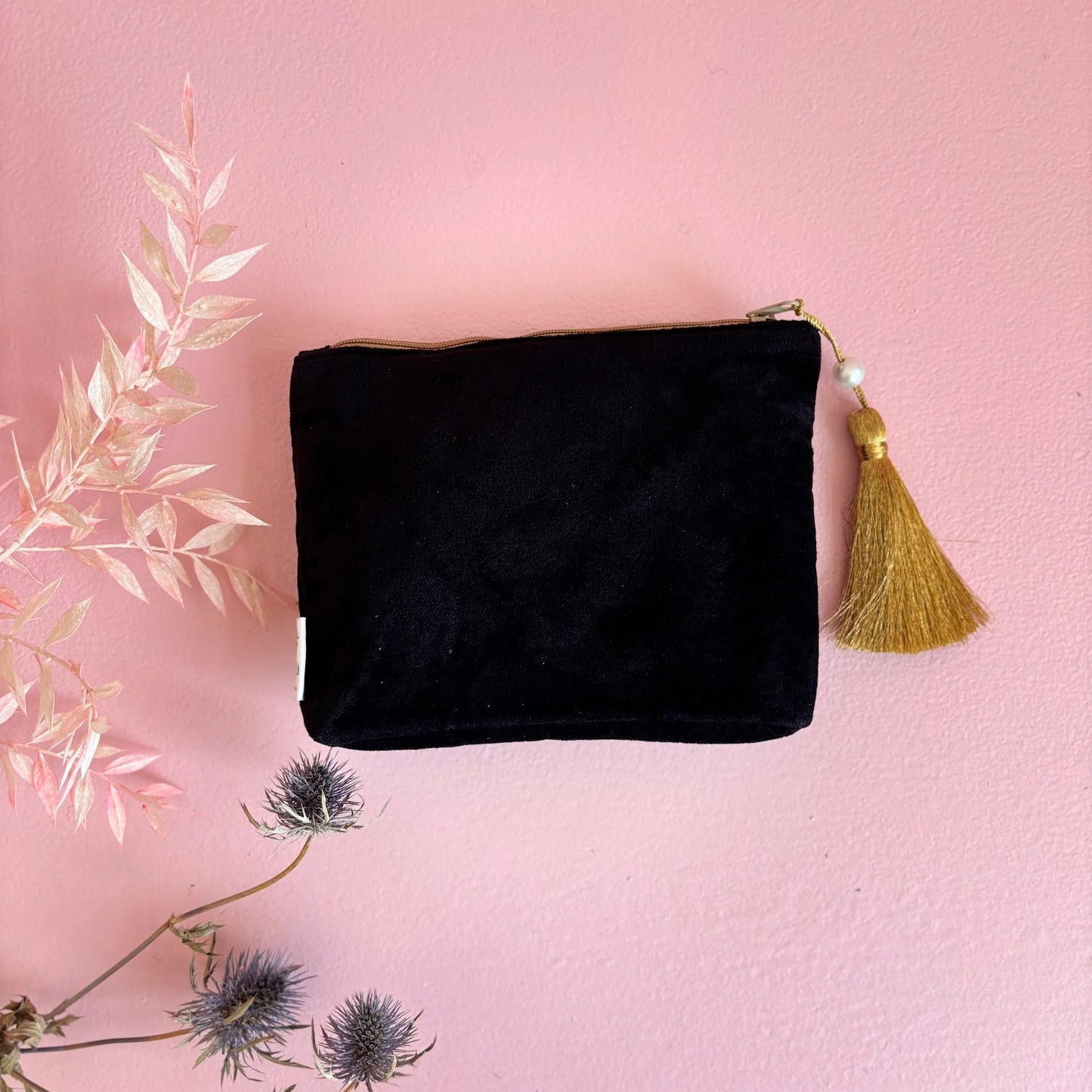 A black velvet vegan-friendly cosmetic pouch with a gold tassel, set on a pink surface beside dried flowers.