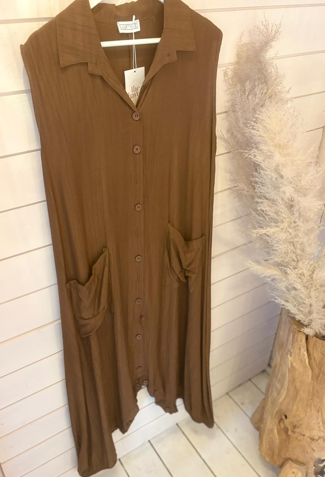 Sleeveless brown jumpsuit dress with a long silhouette and two front pockets, shown on a wooden hanger against a white shiplap wall, beside a vase containing pampas grass.