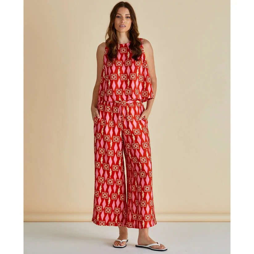 Woman stands against a plain background wearing a sleeveless red patterned top and matching lightweight trousers, hands in pockets, finished with white sandals—an effortless example of ethical fashion.