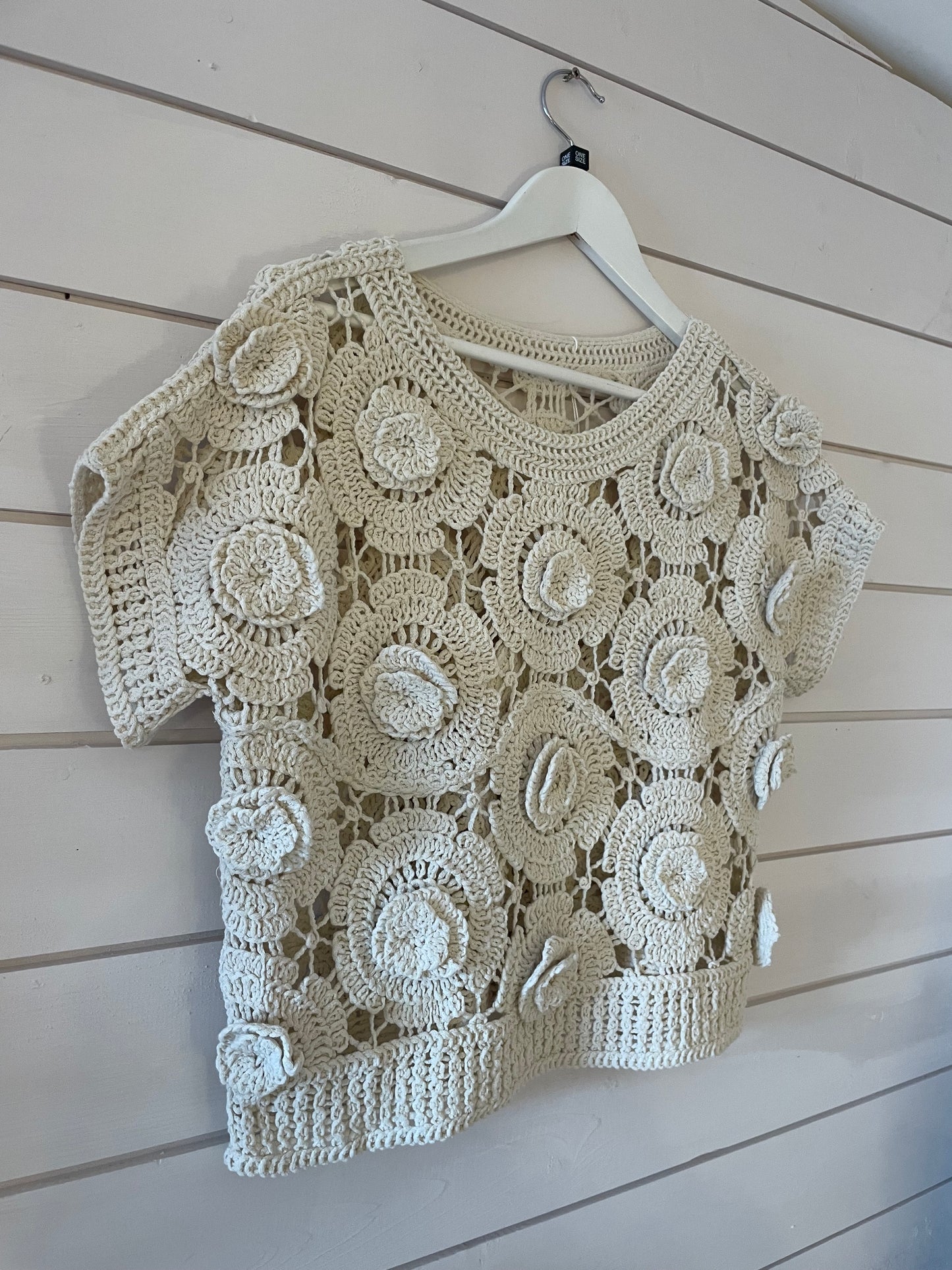 A cream floral jumper with circular flower patterns, this short-sleeved crochet top is an ideal sheer layering piece for versatile spring style. It hangs on a white hanger against a white panelled wall.