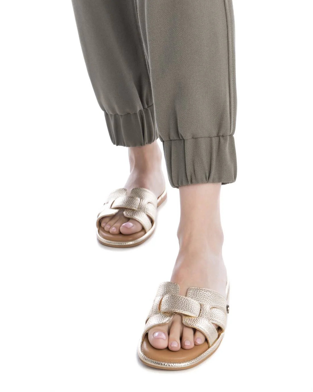 A person wearing olive green cuffed trousers and gold womens sandals, shown from the knees down against a white background.