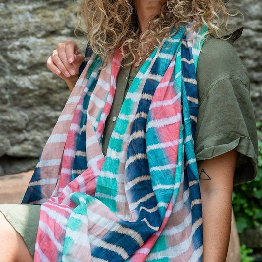 A person with curly hair, wearing a green short-sleeved dress and a lightweight summer scarf, sits outdoors in front of a stone wall.
