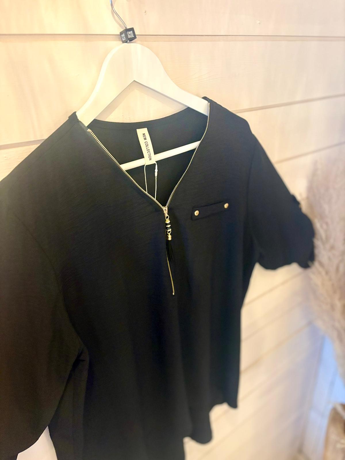 A black short-sleeved blouse with a gold zip and front pocket is shown on a white hanger against a wooden wall.