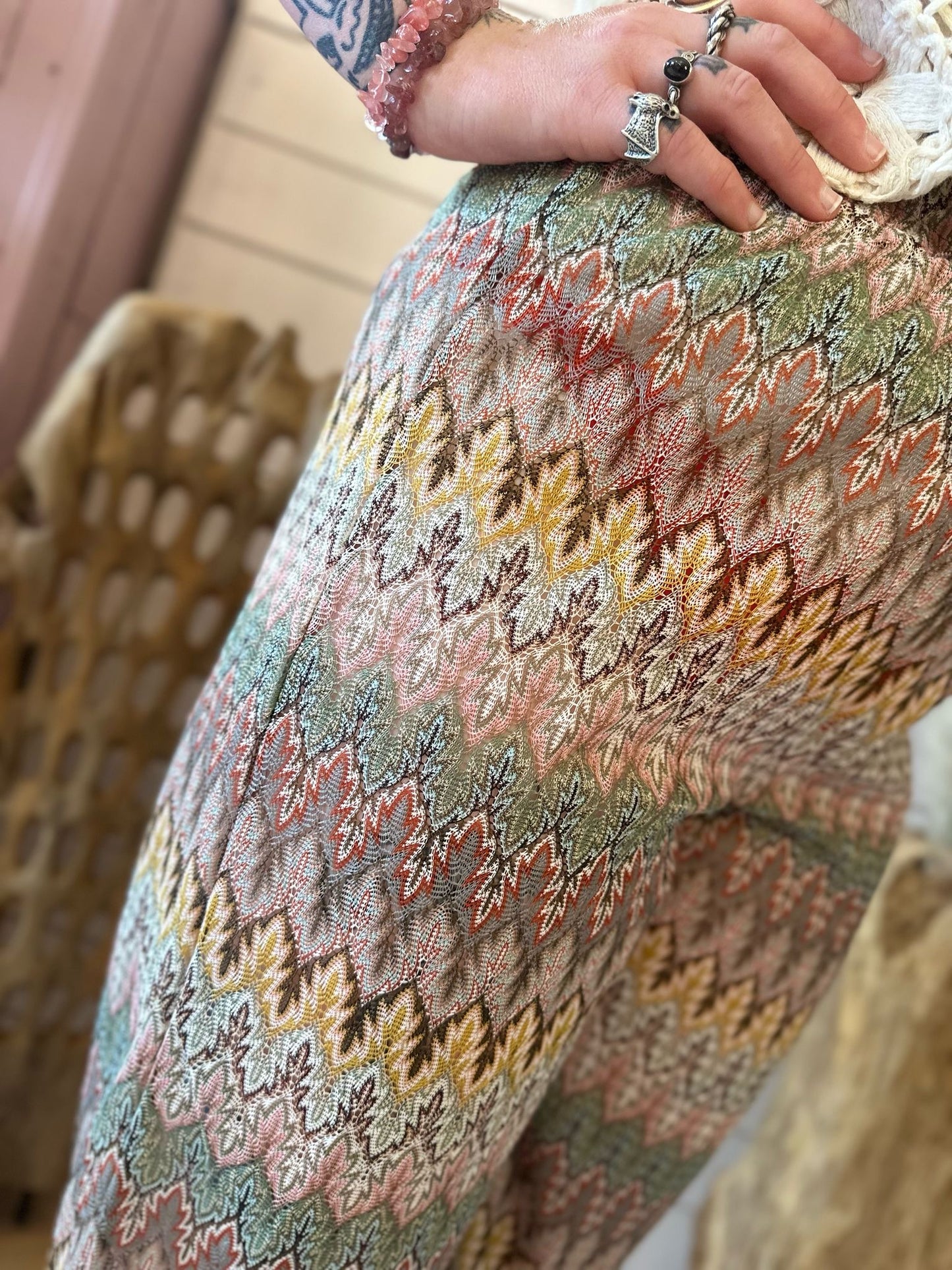 Close-up of a person wearing Aztec Mesh Trousers in earthy tones, featuring multi-coloured patterns, their hand resting on their hip and displaying several rings and a beaded bracelet.