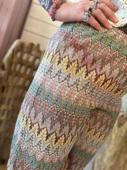 Close-up of a person wearing Aztec Mesh Trousers in earthy tones, featuring multi-coloured patterns, their hand resting on their hip and displaying several rings and a beaded bracelet.