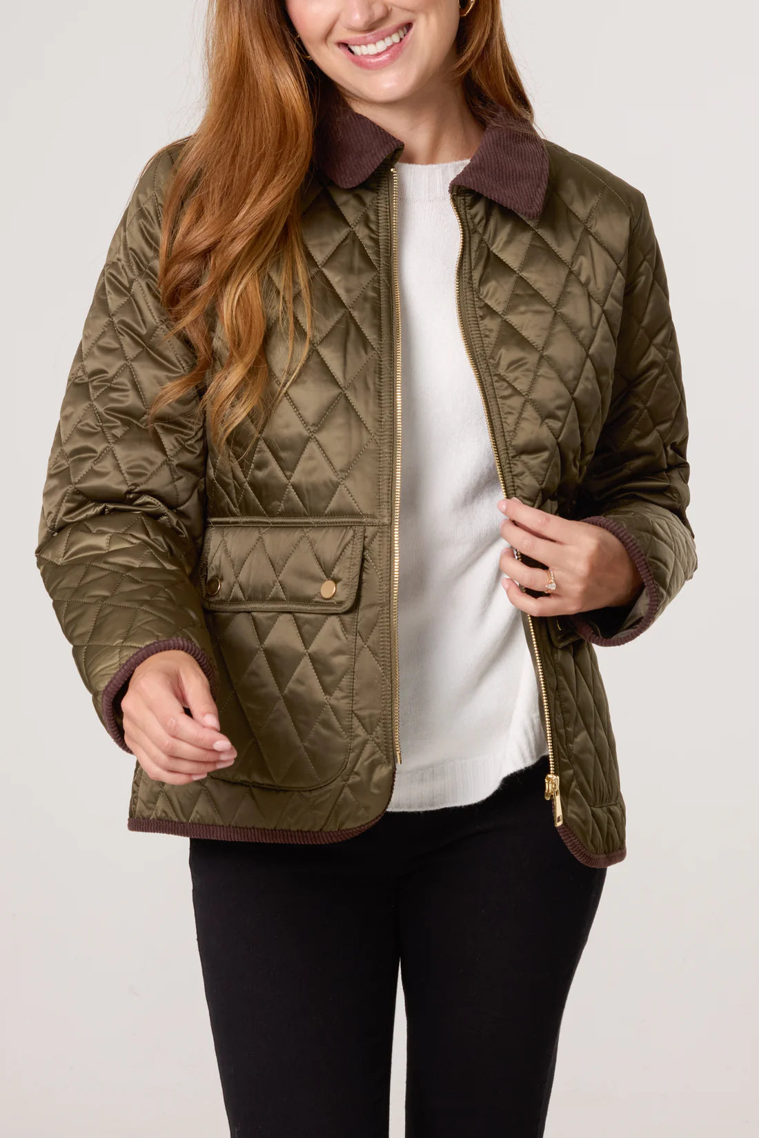 A woman wearing a vegan khaki diamond-quilted coat with brown corduroy collar and cuffs, a white top, and black trousers, zipping up her jacket for extra warmth.