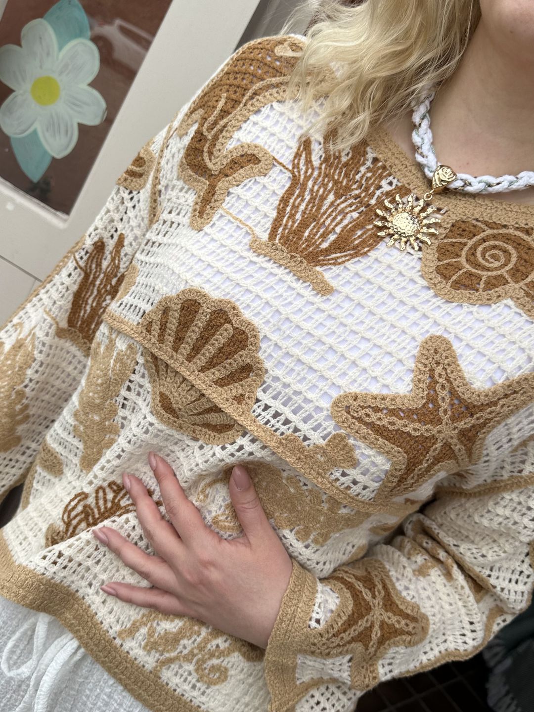 A person is wearing a cream sea life jumper featuring beige and brown seashell and starfish designs. They have a white braided necklace with a gold sun pendant and one hand resting on their torso.