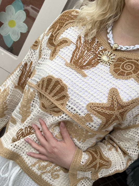 A person is wearing a cream sea life jumper featuring beige and brown seashell and starfish designs. They have a white braided necklace with a gold sun pendant and one hand resting on their torso.