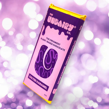 A purple and pink vegan chocolate bar labelled Salted Caramel with UNREAL branding rests on a glittery purple background, showcasing this delicious non-dairy chocolate treat.