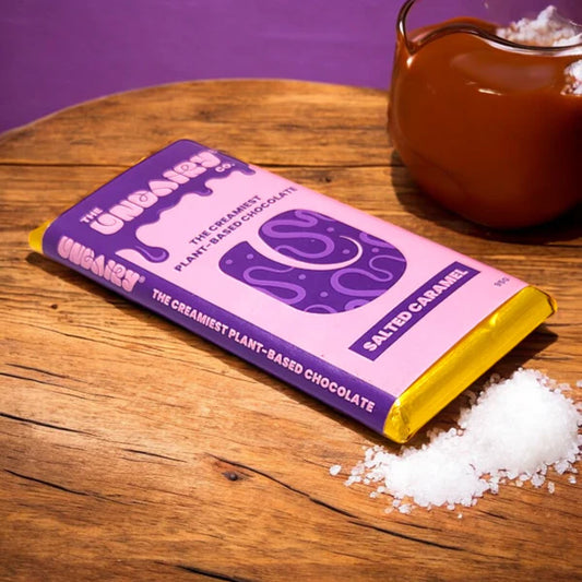 A purple and yellow salted caramel chocolate bar rests on a wooden surface beside coarse salt and a jar of caramel—an irresistible treat for vegan chocolate lovers.