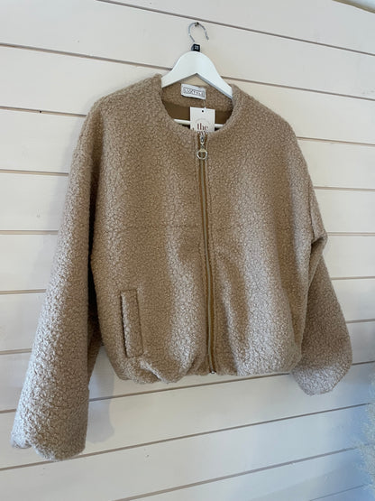 A cream bomber jacket with a relaxed fit, featuring long sleeves, a zip-up front, and two pockets. Made from sherpa and displayed on a white hanger against a white shiplap wall.