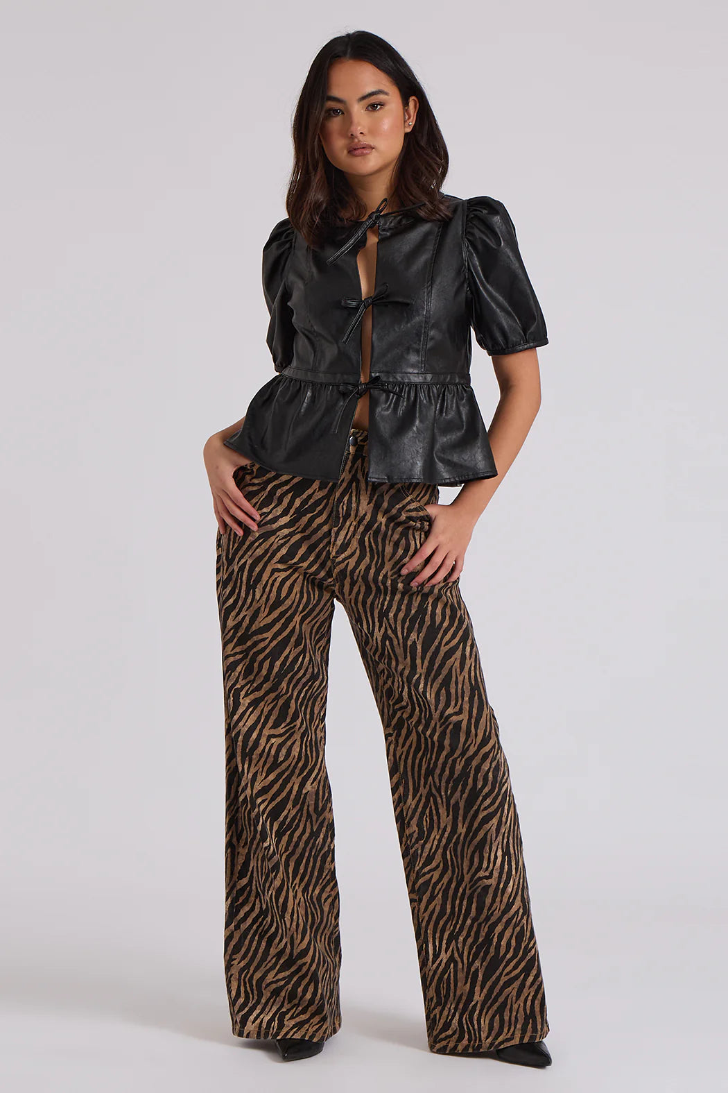 A person wearing a black leather short-sleeved top with a bow and striking Zebra Wide Leg Jeans featuring a flared zebra print pattern stands against a plain background.
