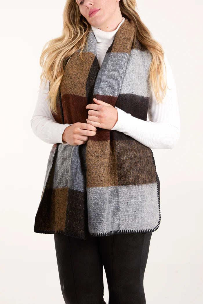 A person wearing a white polo neck jumper and black trousers models a large, cruelty-free, brown, grey, and black checked scarf against a plain background.