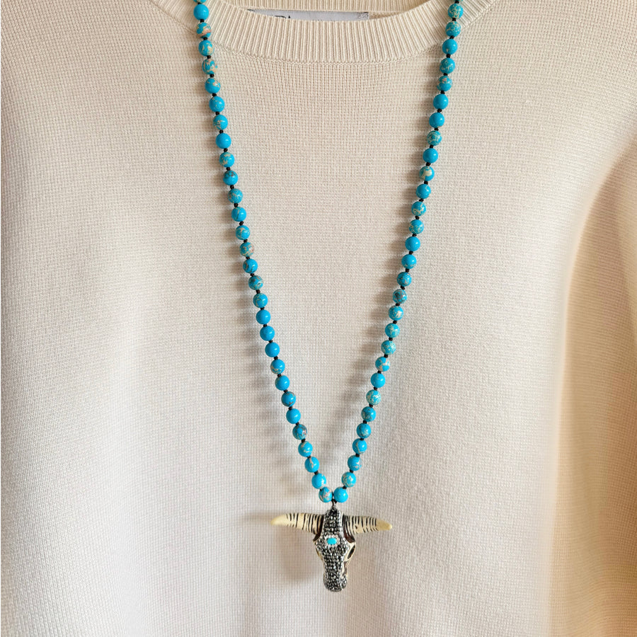 A person wearing a cream-coloured jumper with a green jasper necklace featuring a metallic buffalo head pendant.