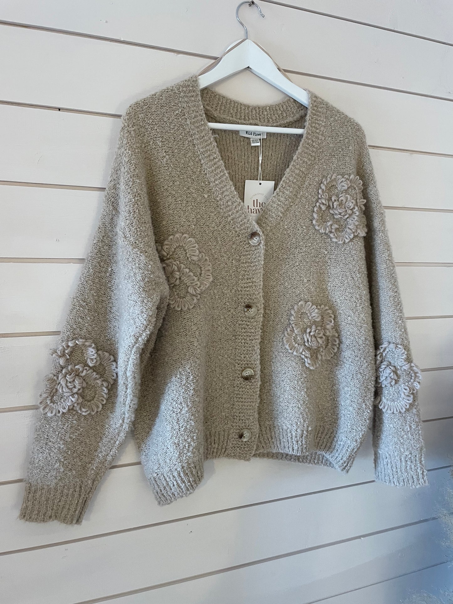 A beige, textured Soft Flower Cardigan featuring large buttons and decorative, raised floral patterns in ultra-soft fabric hangs on a white hanger against a white wood-panelled wall.