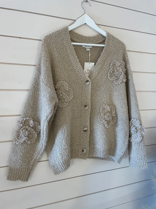 A beige, textured Soft Flower Cardigan featuring large buttons and decorative, raised floral patterns in ultra-soft fabric hangs on a white hanger against a white wood-panelled wall.