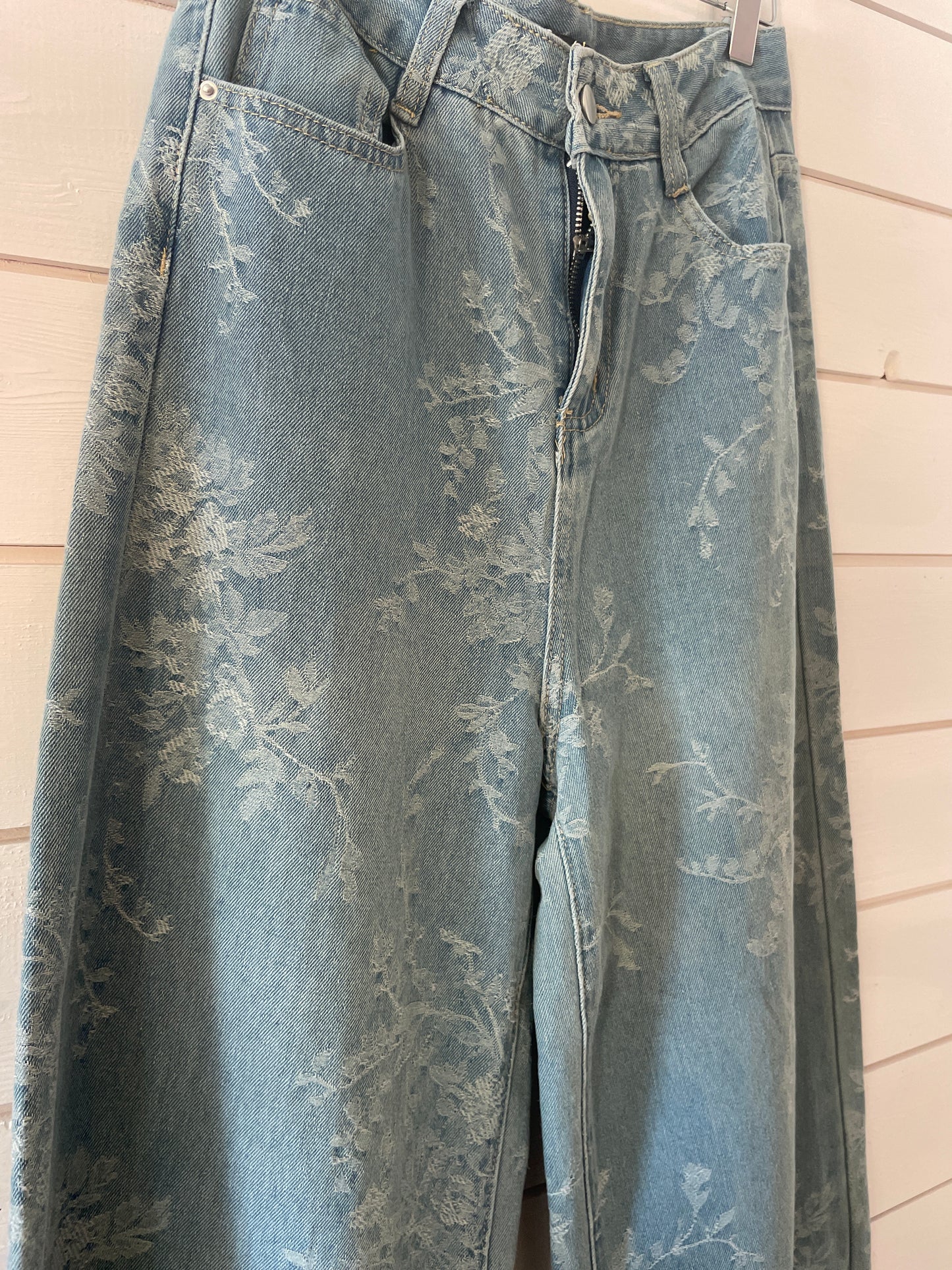 Super soft blue floral jeans with a wide-leg cut hang on a white wooden wall, unzipped at the fly to reveal their subtle pattern.