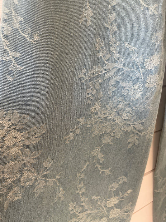Close-up of a light blue curtain featuring a subtle, floral textured pattern—evoking blue flower jeans. The fabric looks soft and slightly shiny, with delicate leaves and flowers woven into the design.