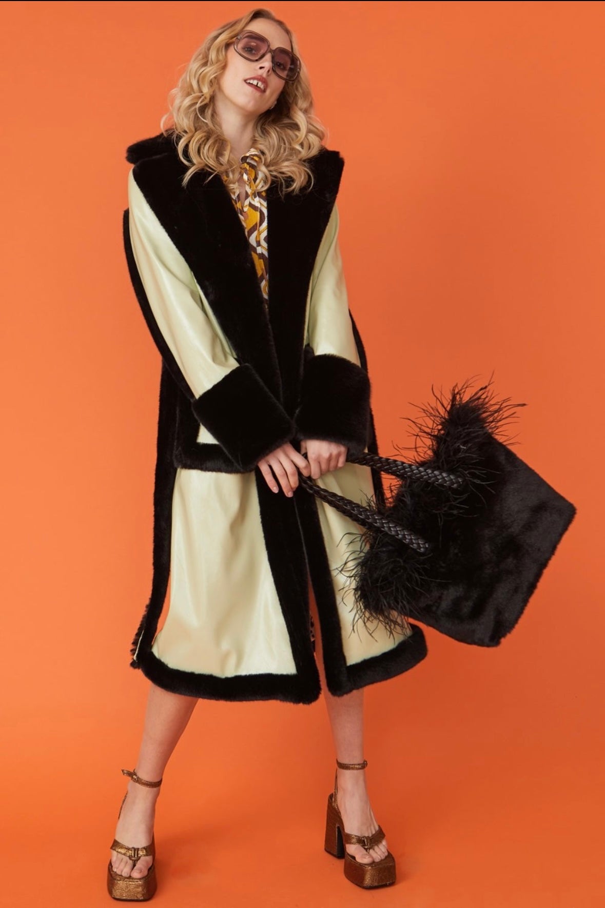 A woman in sunglasses wears a long black and cream faux fur trench coat, a patterned blouse, and platform heels, holding a large black furry bag against an orange background.