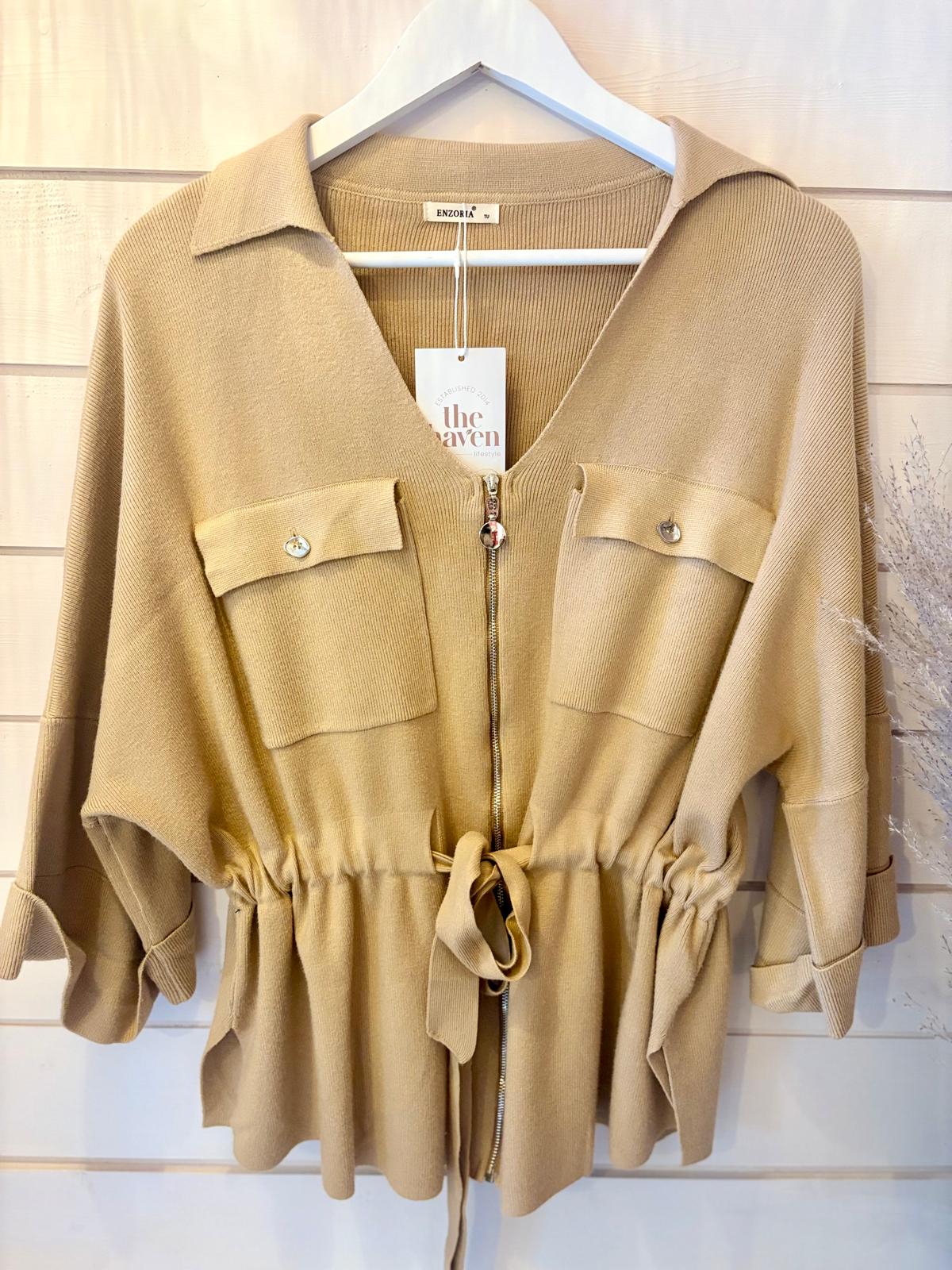 A tan, short-sleeved, collared jacket with a front zip fastening, two chest pockets and a drawstring waist, hanging on a white hanger against a light wood background.