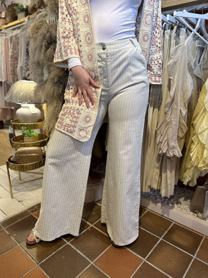 Person wearing pinstripe wide-leg jeans and a white top with a patterned crocheted cardigan, standing in a boutique surrounded by clothing in neutral tones.